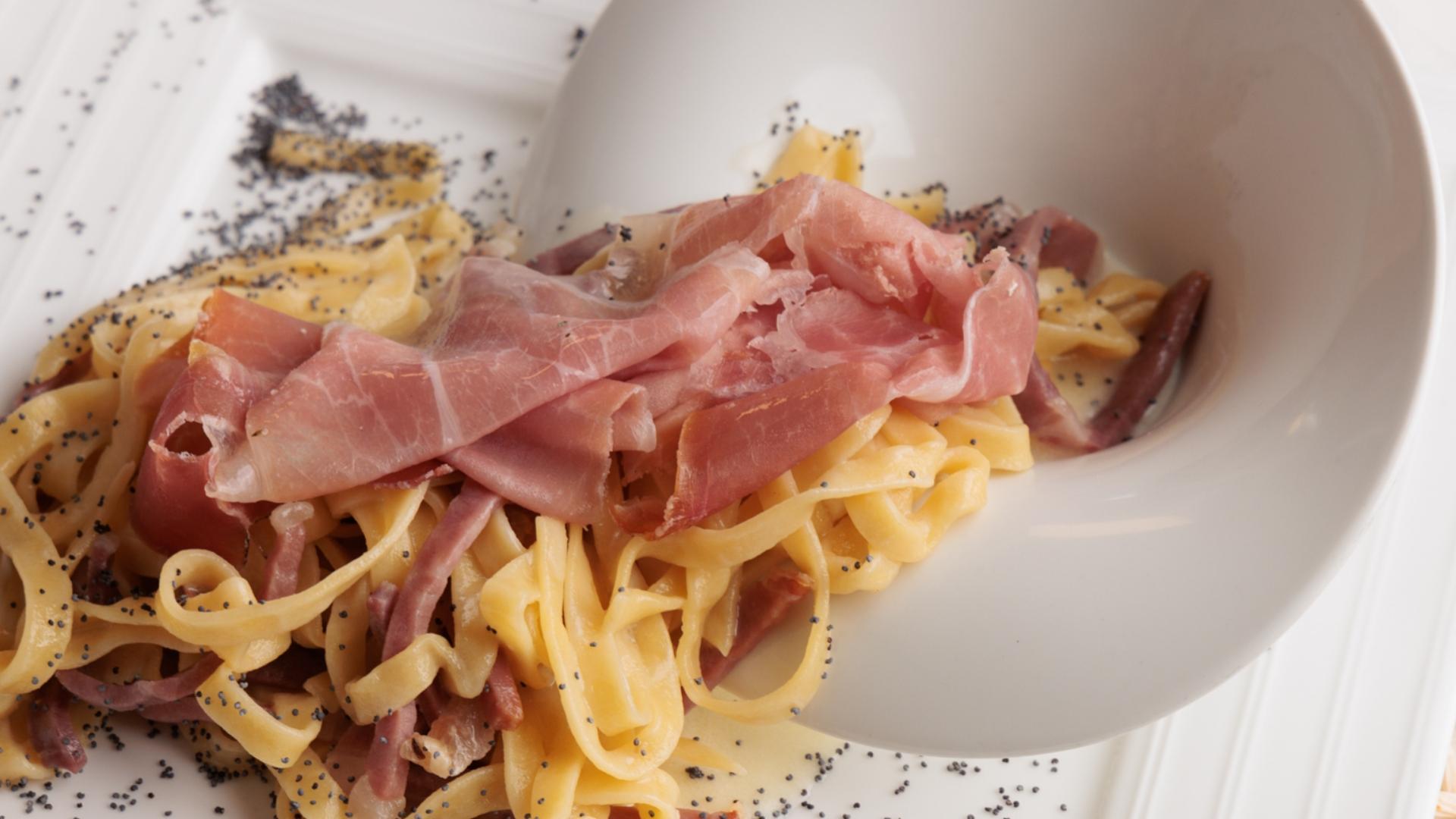 Tagliatelle con prosciutto crudo e semi di papavero su piatto bianco elegante.