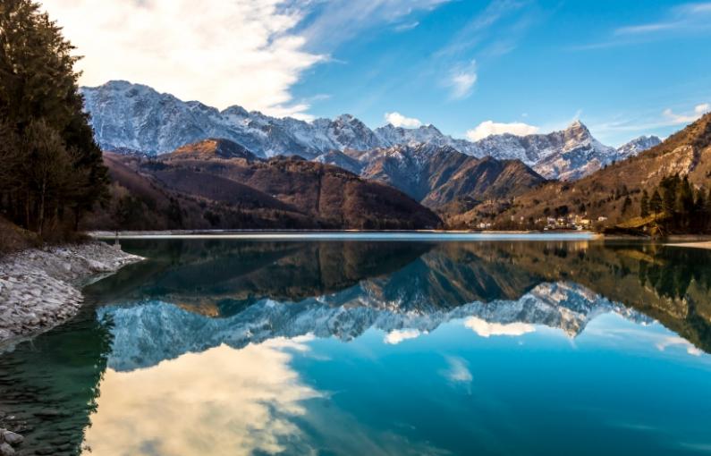 Lago alpino riflette montagne innevate sotto un cielo azzurro e nuvole bianche.